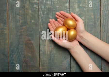 Woman holding shiny golden eggs at light blue wooden table, top view. Space for text Stock Photo