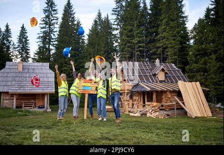 A group of cheerful builders are throwing helmets while posing for a photo at cottage construction site at the forest on a beautiful day. Workers, con Stock Photo