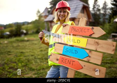 Young female construction worker cheering over wooden table. Construction worker working on wooden house skeleton frame. Construction industry, real e Stock Photo