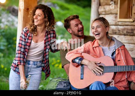 Three cheerful young friends enjoying vacation day and playing guitar in front of wooden cabin. Beuatiful moments. Vacation, holiday, togetherness con Stock Photo