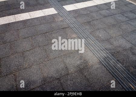 Textured walkway for blind people. Black tactile paving for the visually impaired on the sidewalk. Stock Photo