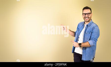 Portrait of cheerful charming mid adult man in denim jacket pointing fingers away at copy space while standing isolated over yellow beige bright backg Stock Photo