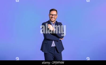 Portrait of confident young adult handsome CEO dressed in blue suit wearing eyeglasses and posing cheerfully against blue background Stock Photo