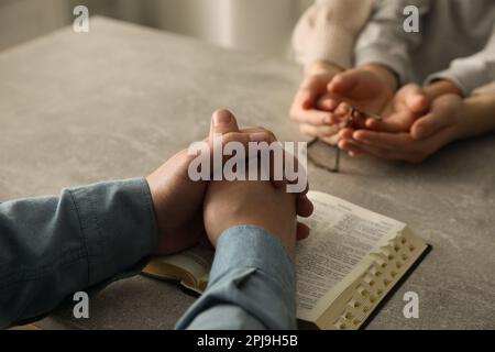 Boy praying and reading Bible with his godparents at grey table indoors, closeup Stock Photo