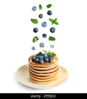 Fresh blueberries and mint leaves falling onto stacked pancakes against white background Stock Photo