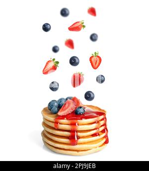 Fresh strawberries and blueberries falling onto stacked pancakes against white background Stock Photo