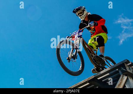 male rider on downhill bike riding wooden drop, background blue sky, racing DH mountain bike, extreme sport games Stock Photo
