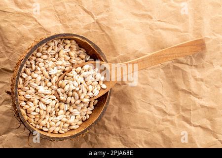 Organic pearl barley in coconut shell with wooden spoon on kraft paper, close-up, top view. Stock Photo