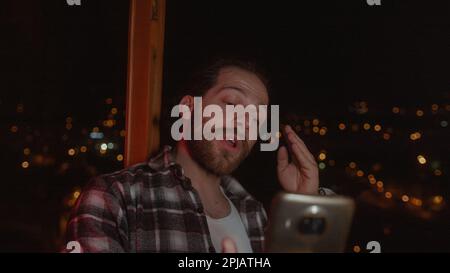Bearded, long-haired handsome young man having a video call in a stunning house with a smart phone; discussing something with someone Stock Photo