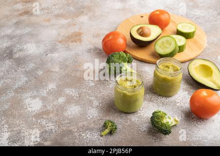 Baby puree with vegetable mix, broccoli, tomatoes, cucumber, avocado infant formula in glass jar on brown concrete background. Side view, copy space, Stock Photo
