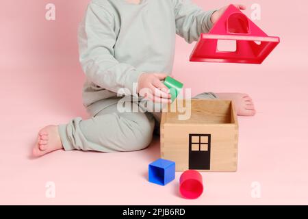 Happy toddler baby plays sorter with geometric shapes on studio pink background. Child boy plays educational toys. Kid age one year eight months Stock Photo