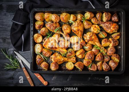 Baked hot chicken legs with spicy potatoes in baking tray on black background Stock Photo