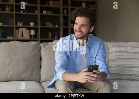 Happy attractive young guy holding cell, sitting on cozy couch Stock Photo