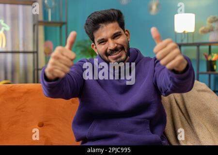 Like. Happy excited indian man looking approvingly at camera showing thumbs up, like sign positive something, good great news, positive feedback. Young hindu guy sitting on couch at home living room Stock Photo