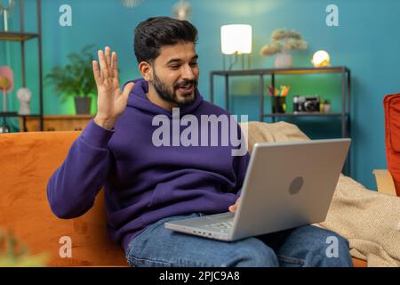 Indian man sitting on couch looking at camera, making video webcam conference call with friends or family, enjoying pleasant conversation. Young hindu guy laughing waving hello at home room Stock Photo