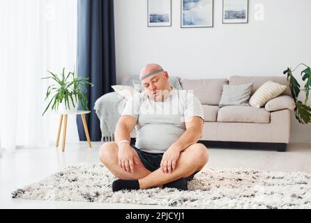 Doing yoga exercises. Funny overweight man in casual clothes is indoors at home Stock Photo