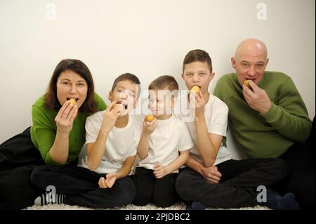 Three brothers three sons father and mother sit on white background eat sweets delicious appetizing little boy peeks what to do white clothes black pants father and mother have green sweaters Stock Photo