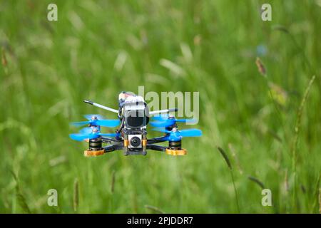Small racing drone also quad copter with blue propellers and a big battery, floats calmly over a beautiful meadow. View directly from the front. Depth Stock Photo