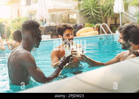 Three young men, one African and two Caucasian, chat and toast with refreshing soft drinks while enjoying a summer day in a swimming pool. Stock Photo