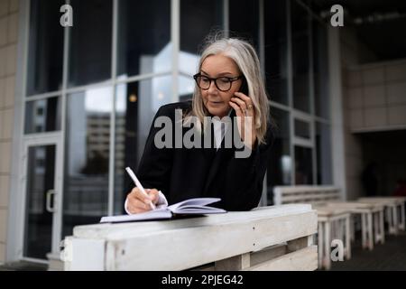 caucasian senior businesswoman taking notes talking on the phone outside the office Stock Photo
