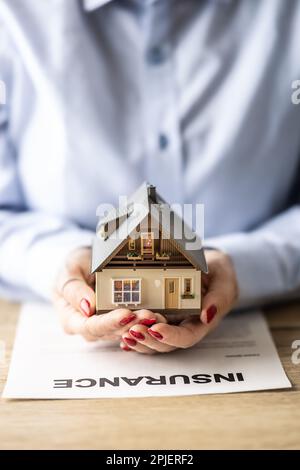 House insurance concept displayed via female hands holding a model of a building on top of an insurance agreement. Stock Photo