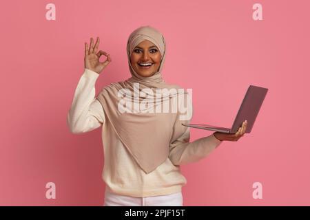 Happy Muslim Woman In Hijab Holding Laptop And Gesturing Ok Stock Photo