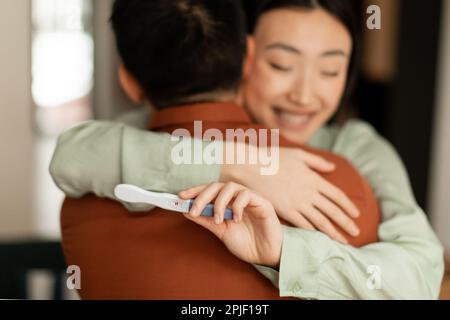 Happy young asian woman holding positive pregnancy test and hugging husband, loving family embracing at home, closeup Stock Photo