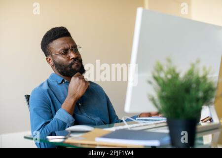 Thoughtful black businessman sitting at workplace and looking at computer screen, thinking about problem solution Stock Photo
