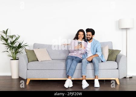 Relaxed multicultural couple sitting on sofa, using smartphone Stock Photo