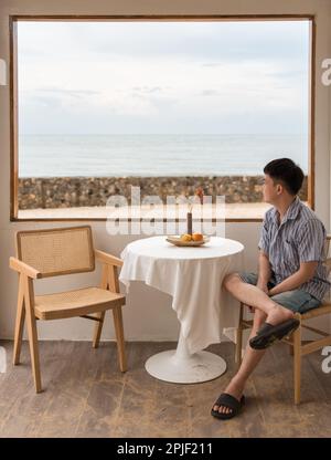 Young asian man sitting and looking at the beach from the window in coffee shop on weekend Stock Photo