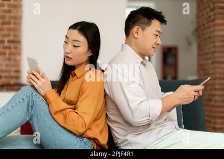 Marital crisis, indifference. Korean spouses using cellphones, sitting back-to-back on sofa, indifferent to each other Stock Photo
