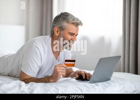 Man chilling on bed, using laptop and bank card Stock Photo
