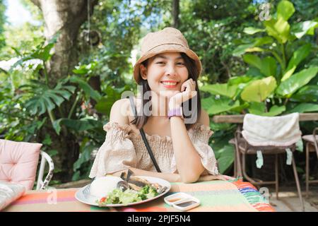 Young asian woman wear hat smiling and eating lunch with fish meat in tropical garden on weekend Stock Photo