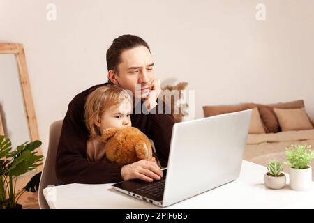 A man works at home on a laptop. The child distracts from work, misses. The father and his daughter are sitting at the computer. Two-year-old girl hug Stock Photo
