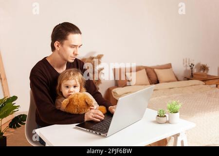 A man works at home on a laptop. The child distracts from work, misses. The father and his daughter are sitting at the computer. Two-year-old girl hug Stock Photo