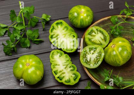 Parsley sprigs, green tomatoes in a plate and on the table. Dark wooden background. Stock Photo