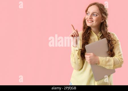 Beautiful redhead woman with laptop pointing at something on pink background Stock Photo