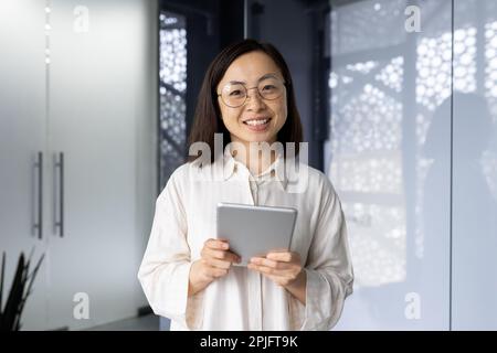 Portrait of successful asian female programmer inside office, young woman smiling and looking at camera, using tablet computer to test application software, workplace inside office. Stock Photo