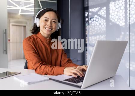Young successful female programmer at workplace inside office working with laptop, coding software, smiling and looking at camera, female worker with headphones online music and podcasts. Stock Photo