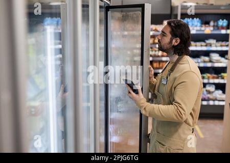 Side view portrait of male worker in supermarket inspecting freezer inventory and holding digital tablet Stock Photo