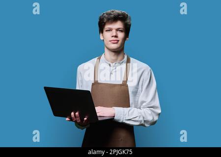 Young male in an apron with laptop, on blue background Stock Photo