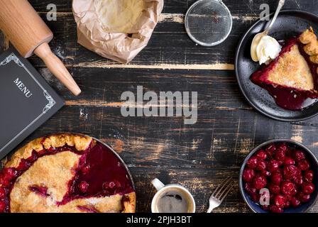 Baking background with sliced cherry pie, flour, rolling pin on the black wooden table. Ingredients for baking/dessert or pie making Stock Photo