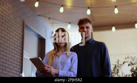 Happy two young colleagues standing in cafe. Lady holding laptop. Couple posing at camera. Lifestyle, partnership, job concept. Real time Stock Photo