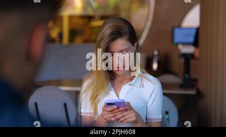 Confused young woman reading message on mobile phone during dating in cafe. Lady sitting in front of a man and looking at smartphone screen Stock Photo