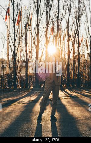 An elegant man in a cream suit and striped tie stands tall, holding a cup. The backlight with the deciduous trees lining creates long, dramatic shadow Stock Photo