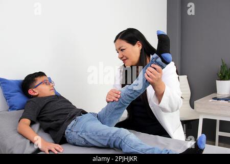 Latina pediatrician woman doctor attends a 7-year-old boy in her office checks his leg, knee and foot for falls and diseases such as rickets, blount Stock Photo