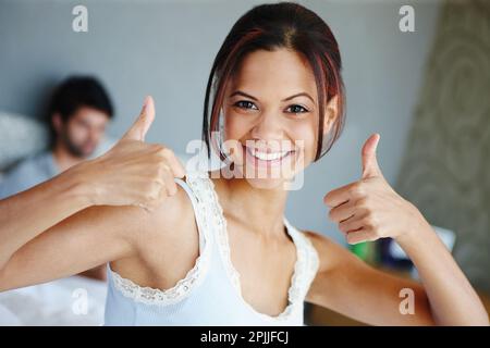 Thumbs up if you had a good nights rest. Portrait of a happy woman with her thumbs up sitting on a bed with her boyfriend in the background. Stock Photo
