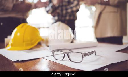 Close up view of engineer worktable with glasses, safety helmet and blueprint on desk in office room. Stock Photo