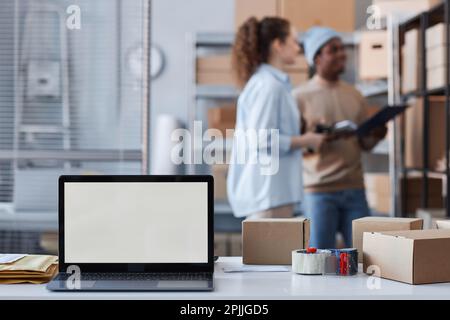 Laptop with white empty screen standing on desk among boxes against two young intercultural managers standing in front of shelf Stock Photo