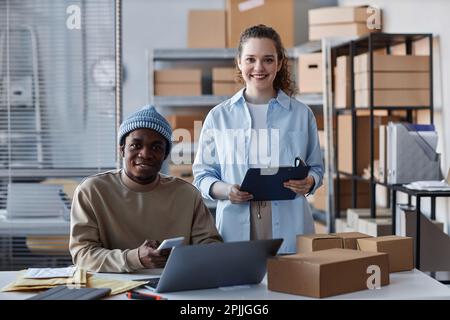 Two happy young intercultural employees in casualwear looking at camera whil.e standing by workplace with laptop and taking new orders Stock Photo
